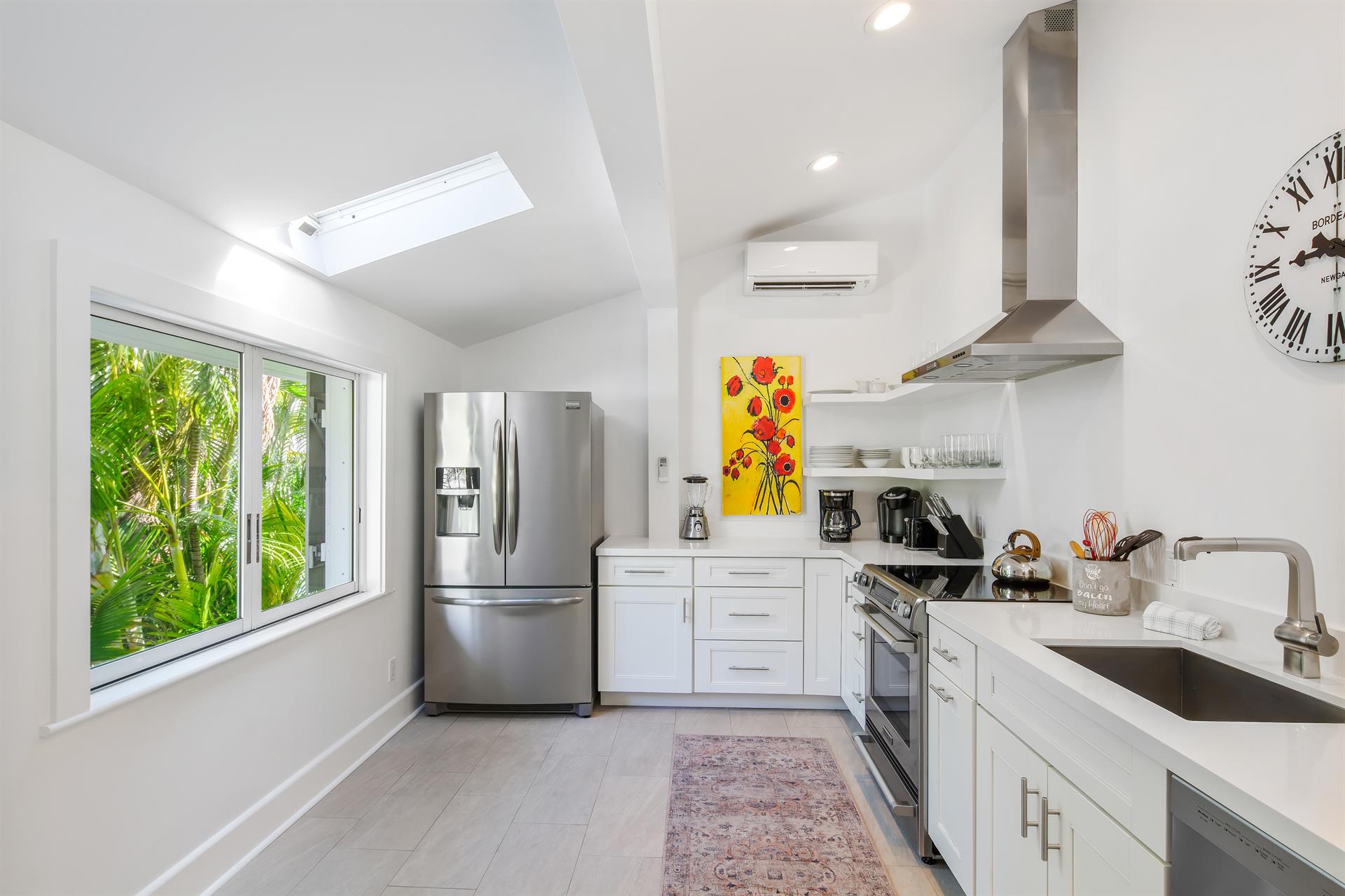 Bright galley kitchen with stainless steel appliances, white cabinetry, skylight, and garden window