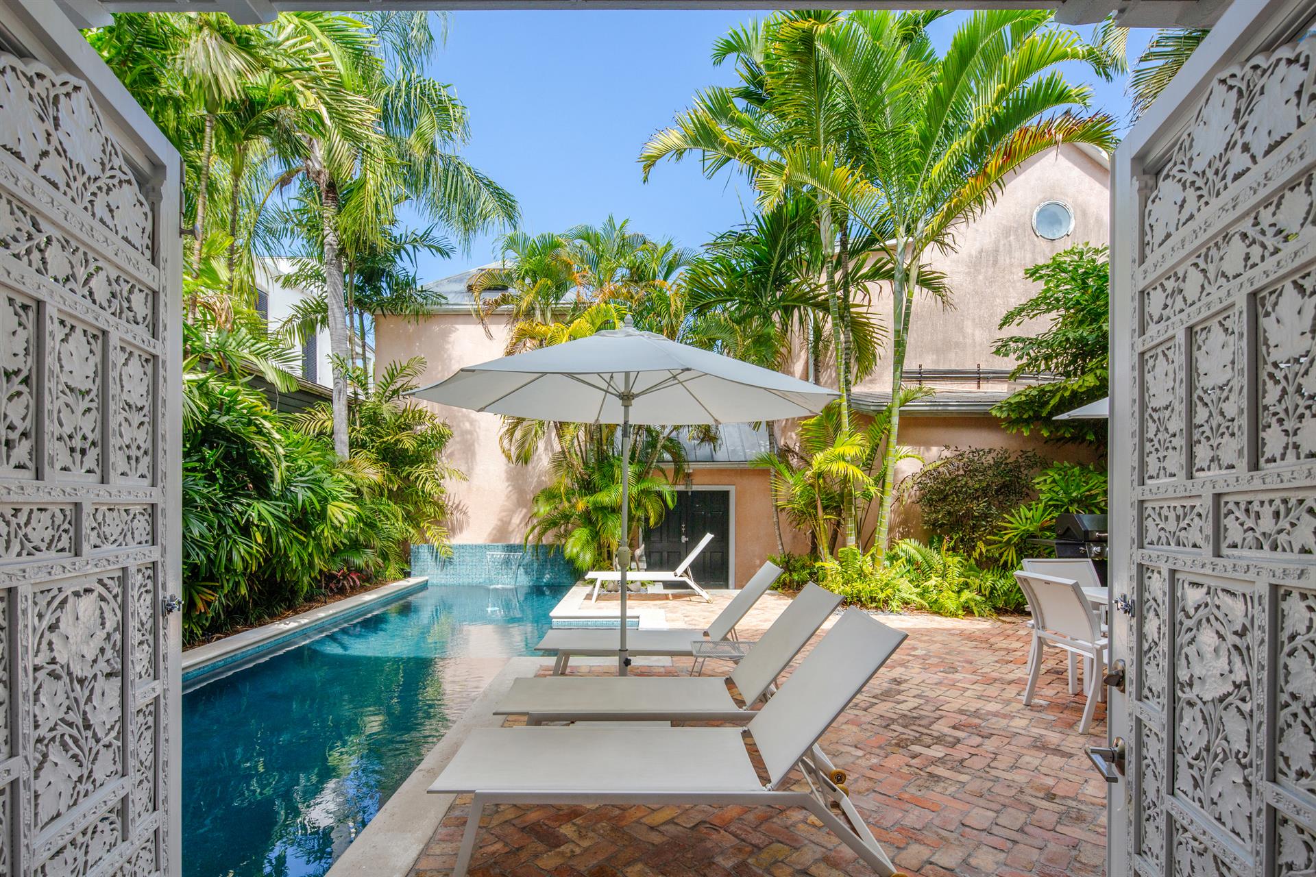 Private courtyard pool viewed through ornate carved white doors, flanked by tropical palms and brick pavers